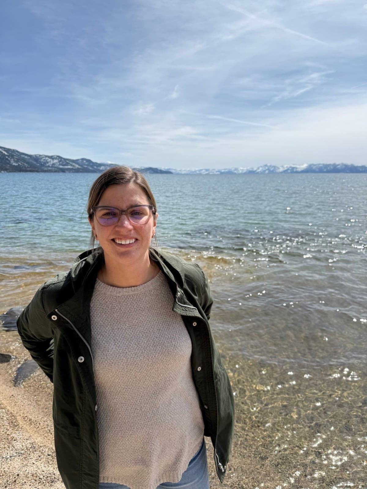 Anita Burke on the shore of Lake Tahoe, snow on the mountains behind her