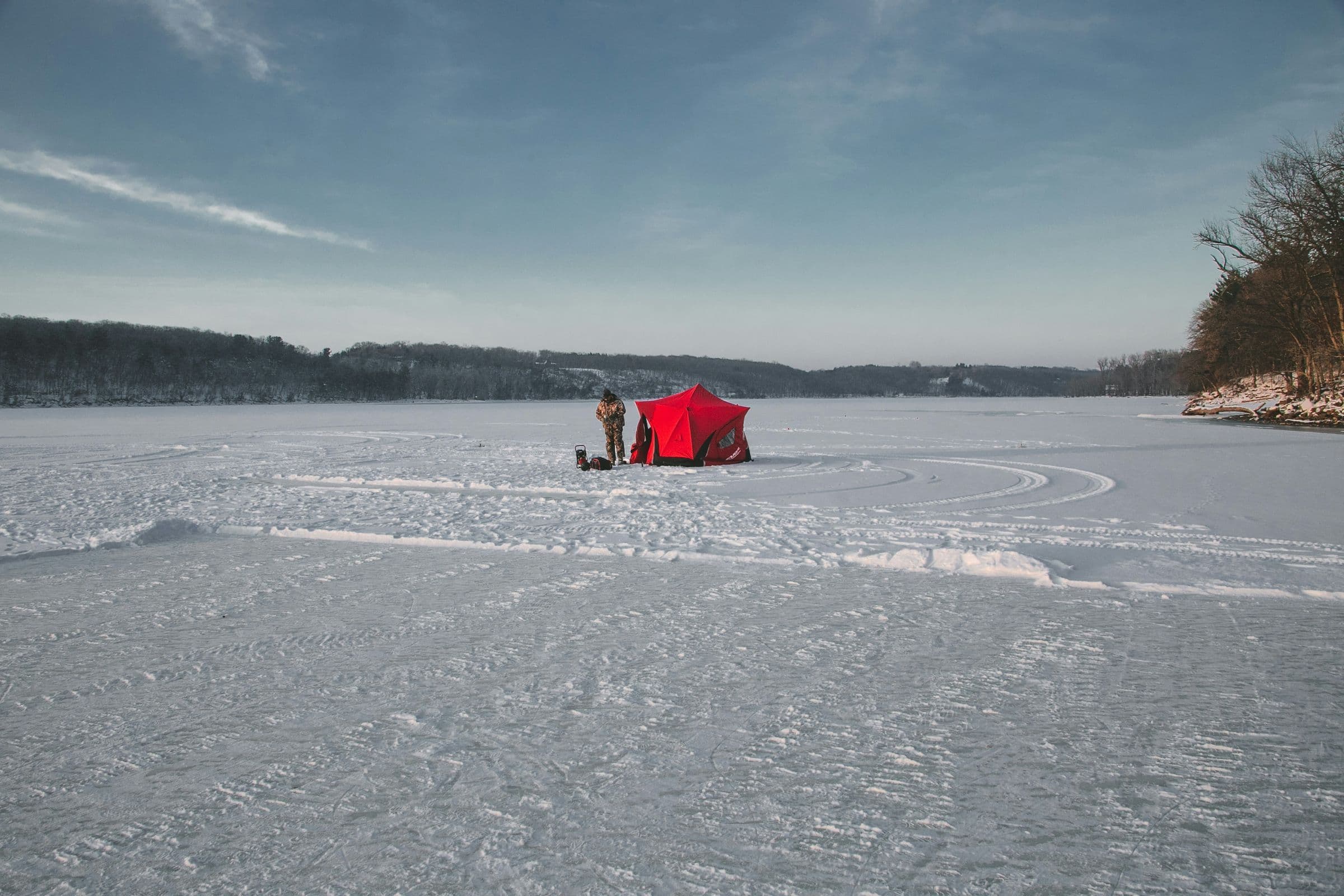 Man Bundled up ice fishing with a red tent on the lake