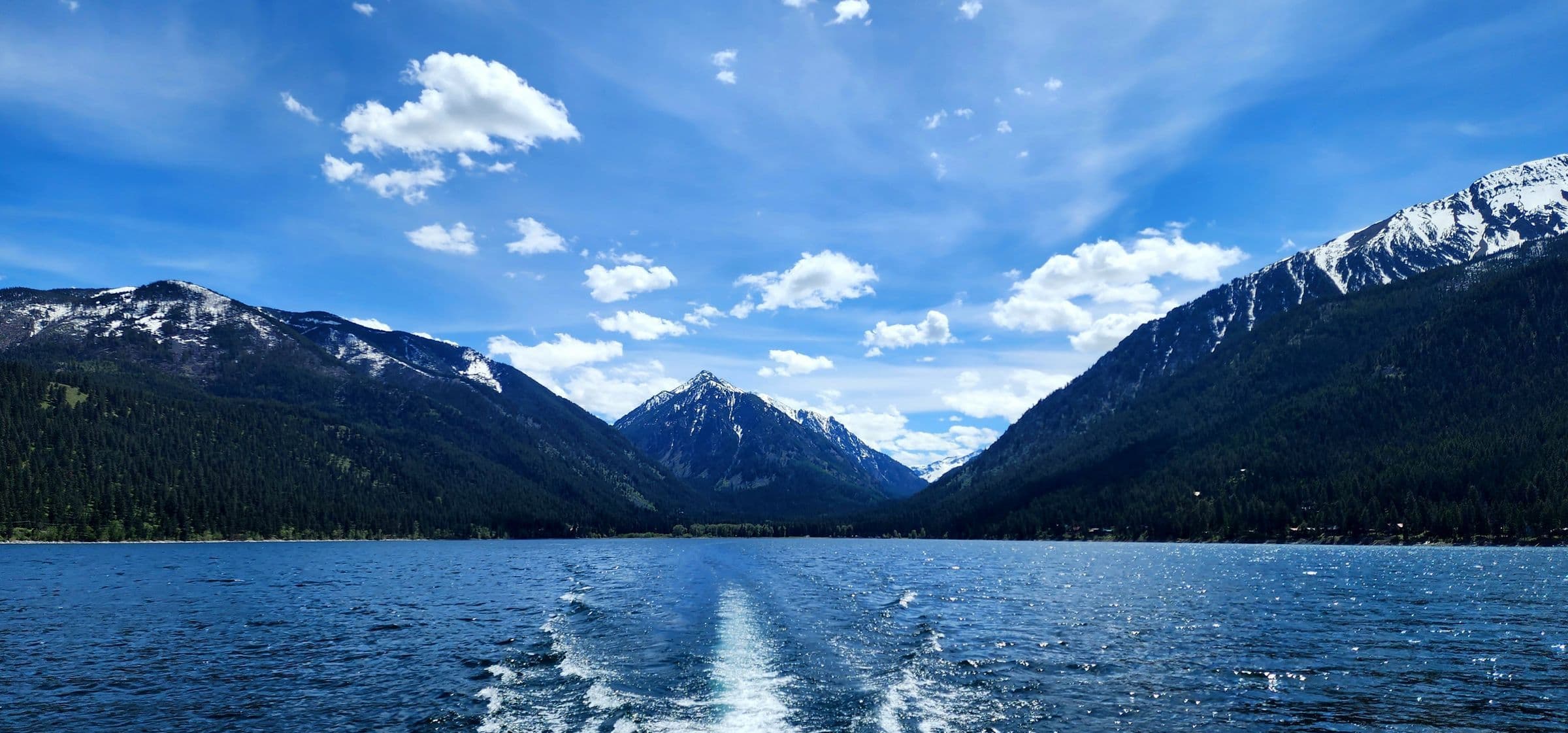 Wallowa Lake clear blue skies and water with wake from behind a boat. Mountains surrounding in the distance
