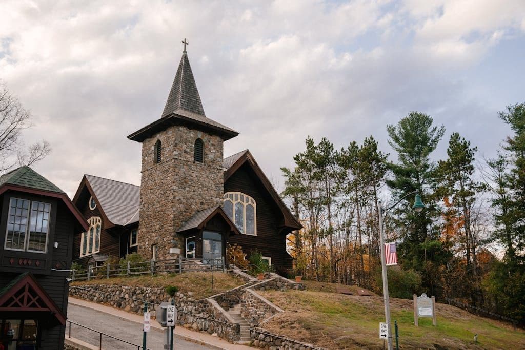 Low angle of ancient St Eustace Episcopal Church with cross on sharp roof located on hill surrounded by autumn trees against cloudy sky in Lake Placid