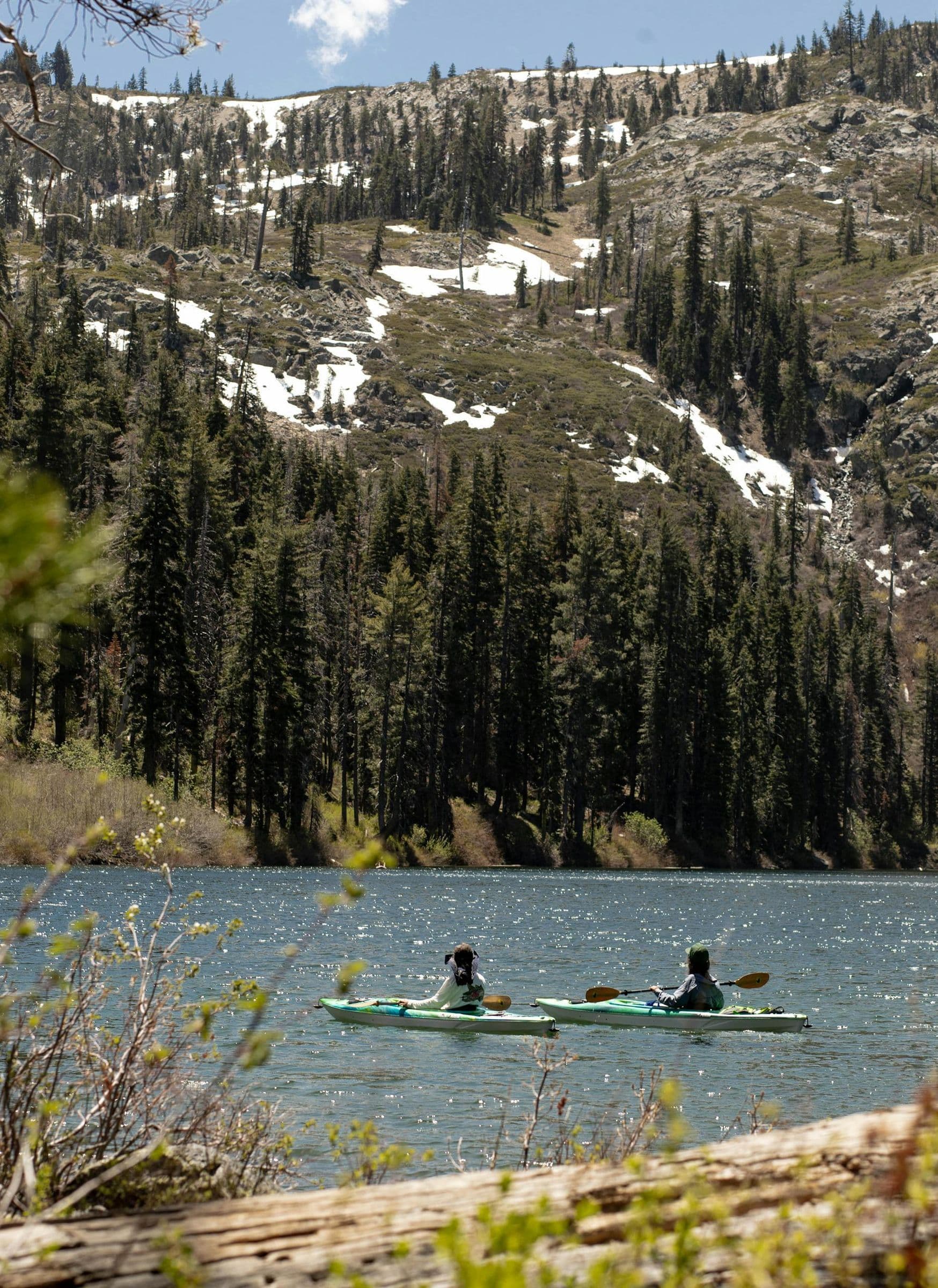 Two people kayaking on a serene mountain lake in California surrounded by lush pine forests.