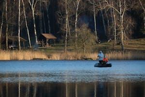 Two people fishing on a tranquil lake surrounded by bare trees in early spring.