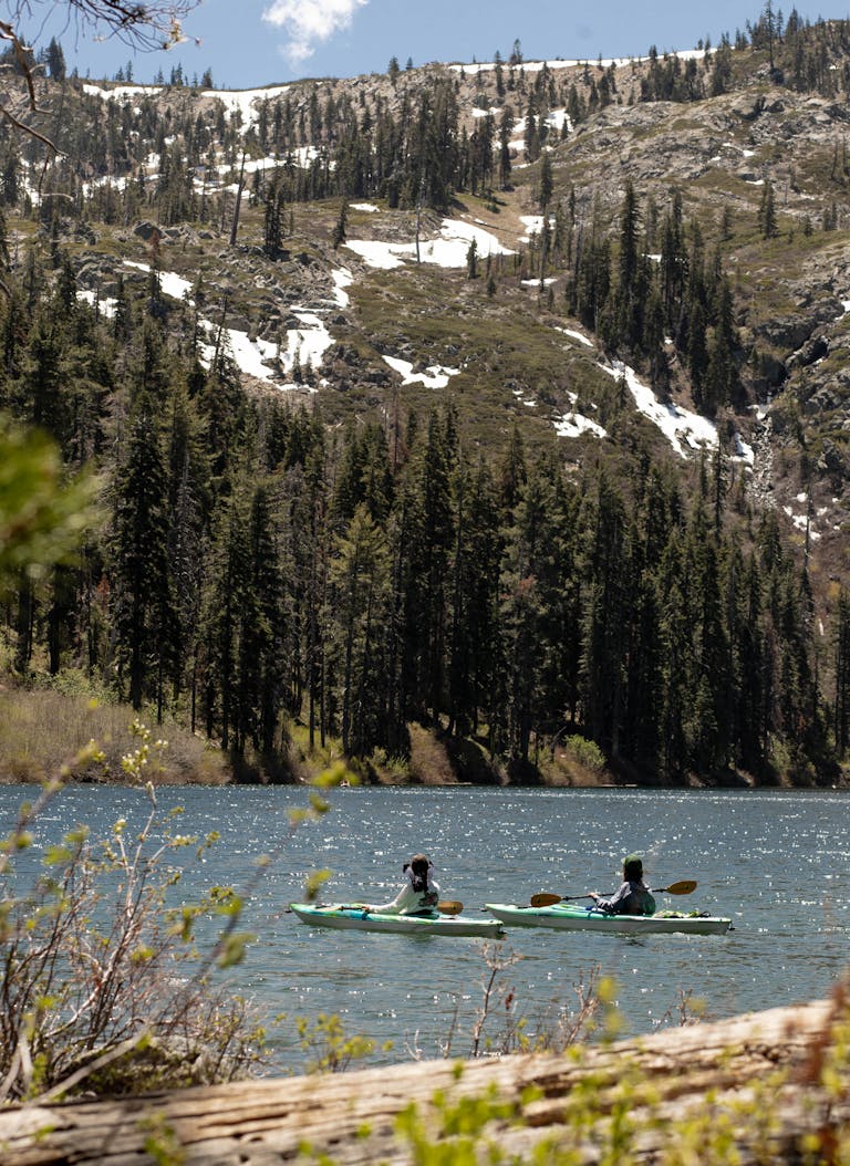 Two people kayaking on a serene mountain lake in California surrounded by lush pine forests.